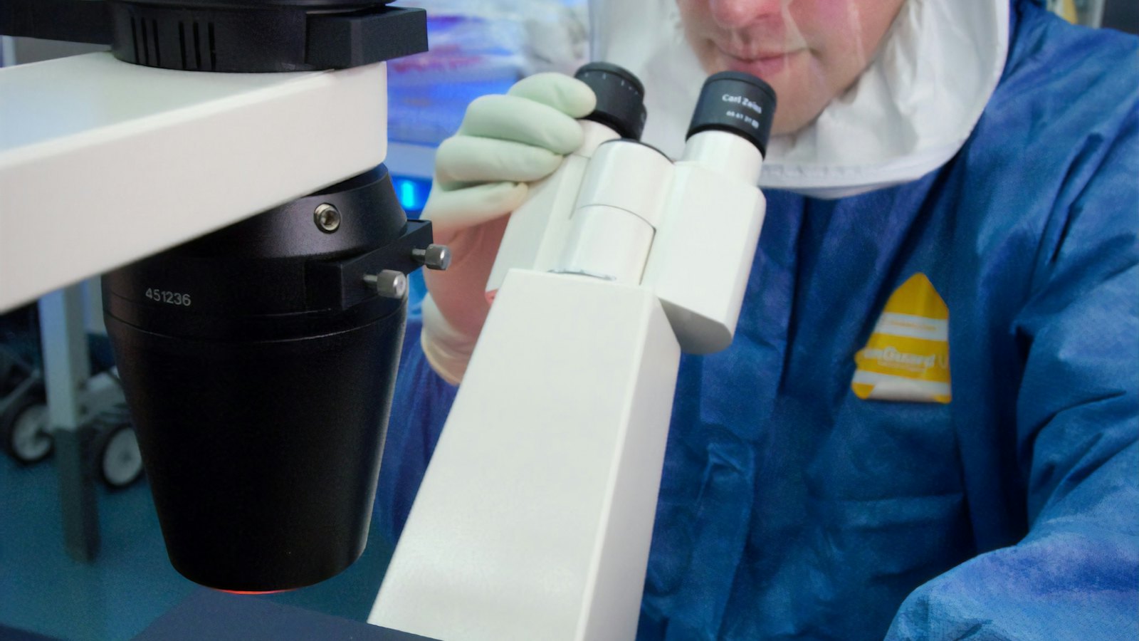 Lab specialist in protective gear looking through a microscope